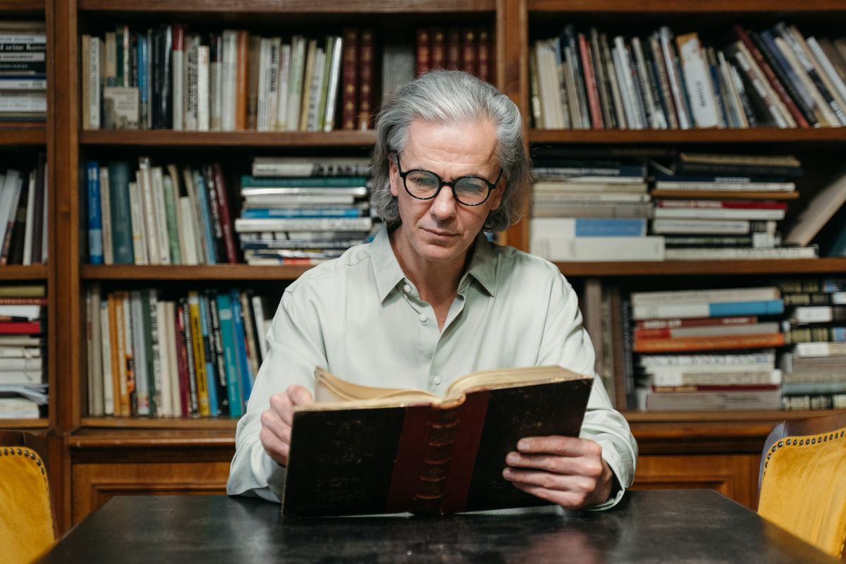 An older adult man with glasses reading a book in front of bookshelves.