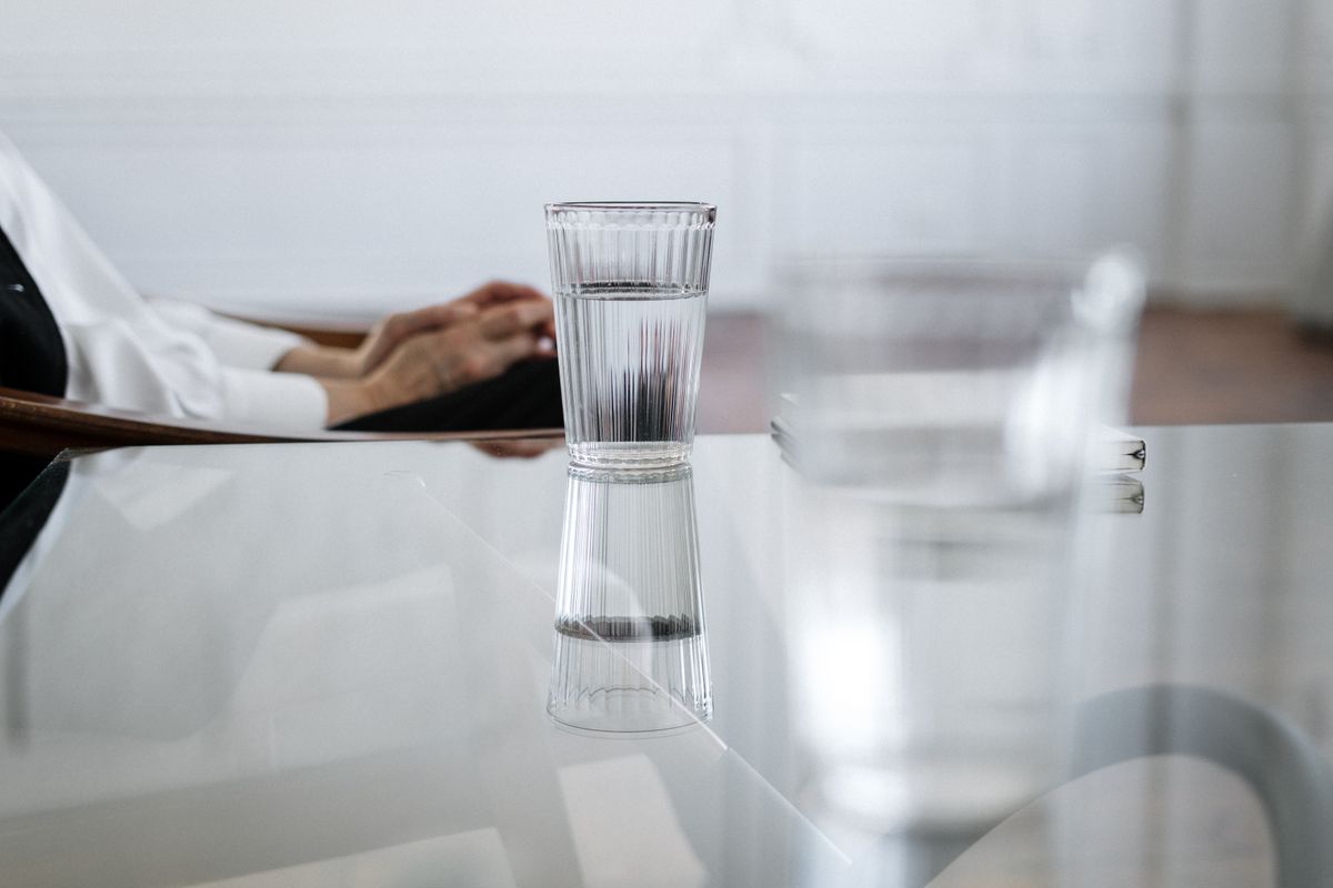 A glass of water on a reflective table, with a seated person's hands in the background.