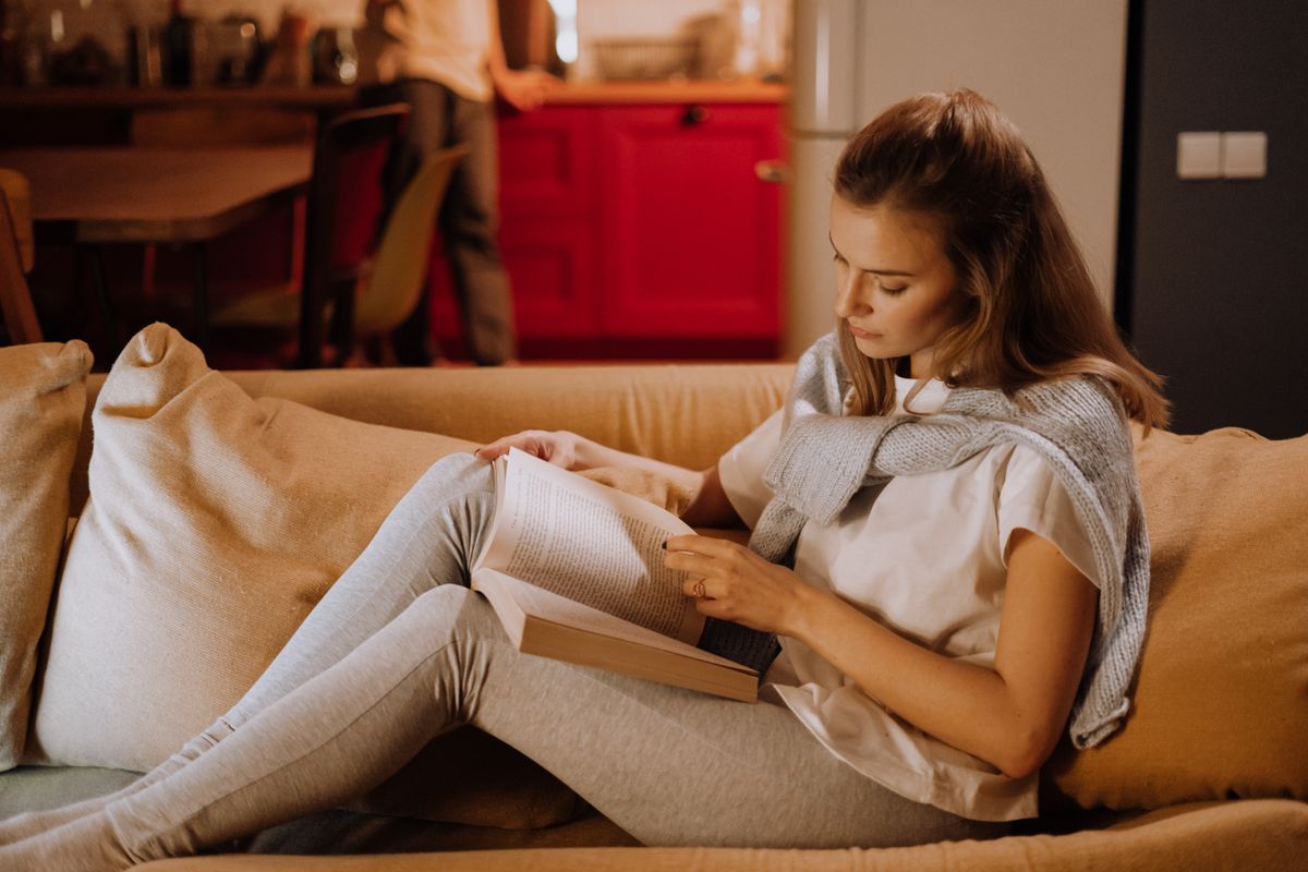 An adult woman seated on a couch reading a book at home.